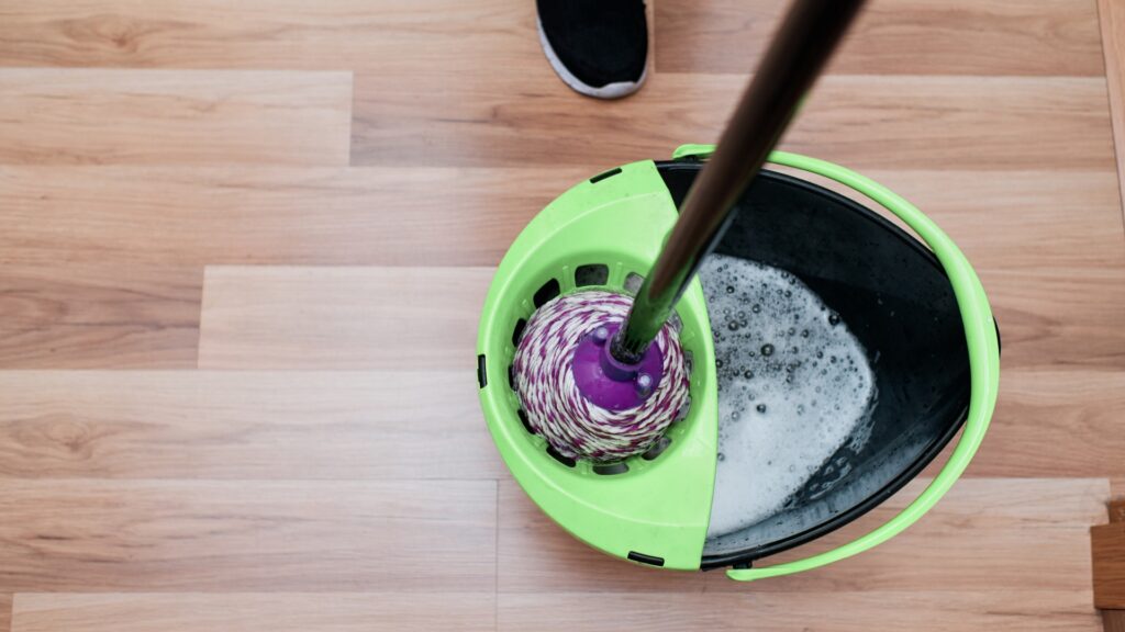 En moppe og spand til gulvvask i brug, med synlige ben af personen, der holder moppen - essentielle redskaber til vedligeholdelse af trægulve.