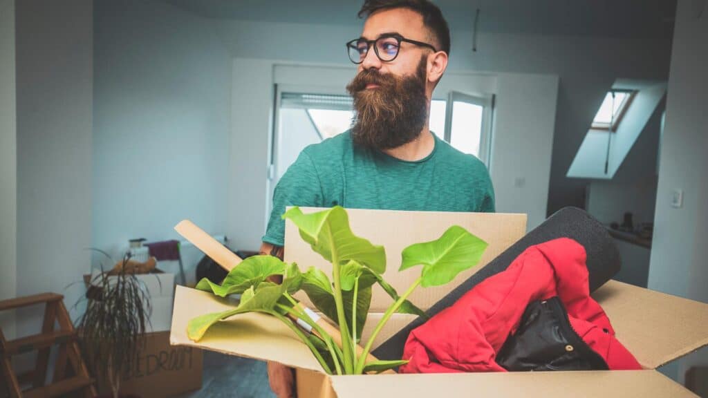 En ung mand med langt skæg og briller holder en kasse med personlige ejendele og friske blomster, klar til fraflytning.